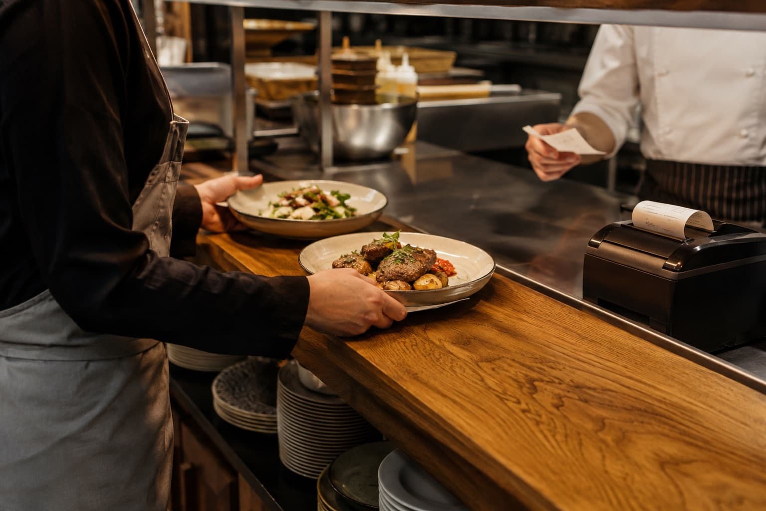 Restaurant team handing prepared plates across a kitchen pass beside a thermal printer